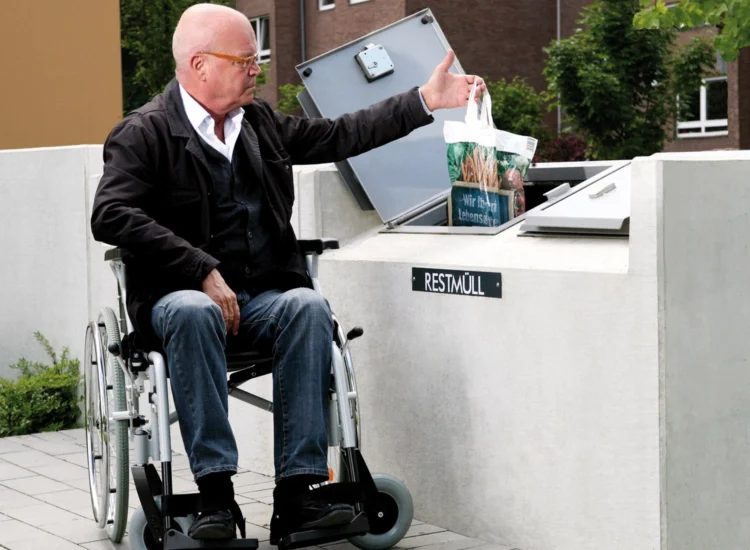 Ein Mann mit Rollstuhl wirft im Sitzen auf Brusthöhe in den tiefergesetzten Müllsammler seinen Müllbeutel.