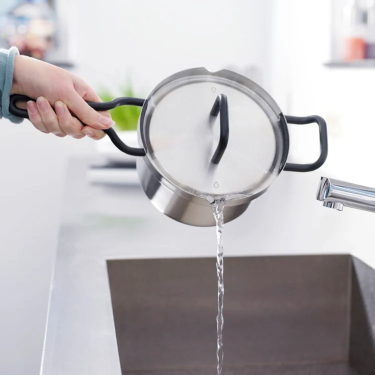 Eine Person gießt Wasser aus dem Topf mit Deckel in ein Spülbecken